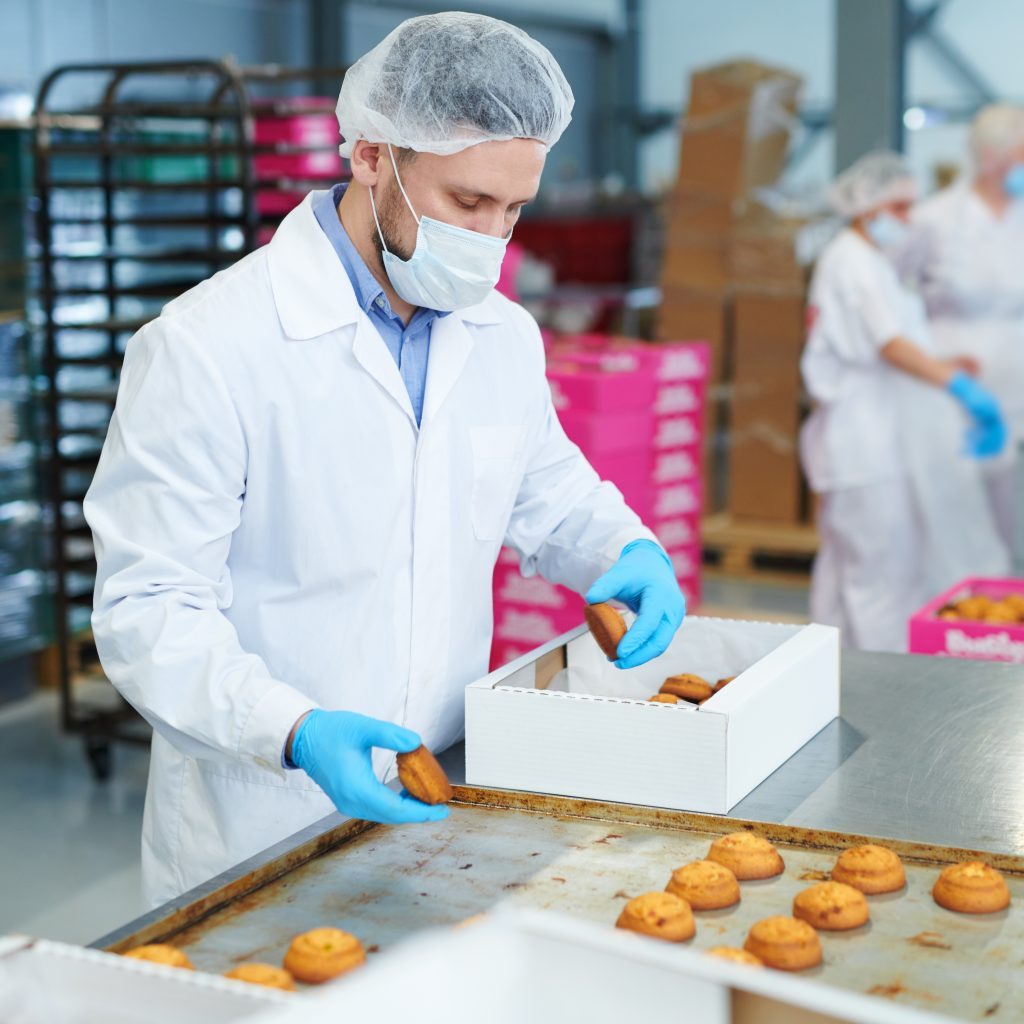 Confectionery factory worker in white coat collecting freshly baked pastry from tray and putting it into paper box.